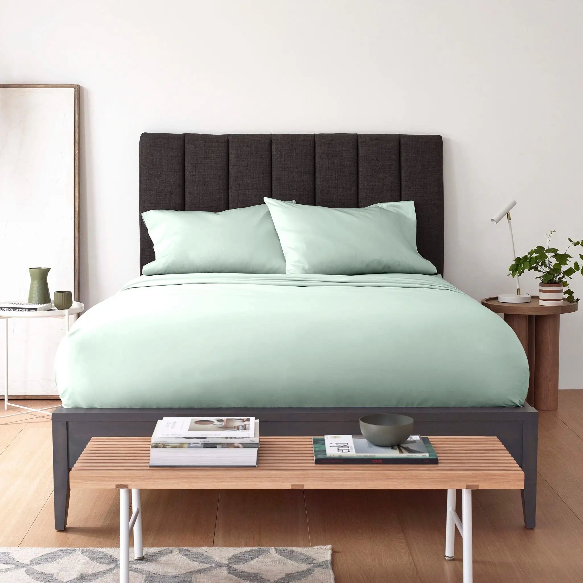 A bed dressed in pale mint green bedding with a dark upholstered headboard and matching dark footboard, styled in a bright bedroom with a bench, side table, lamp, and small plant.