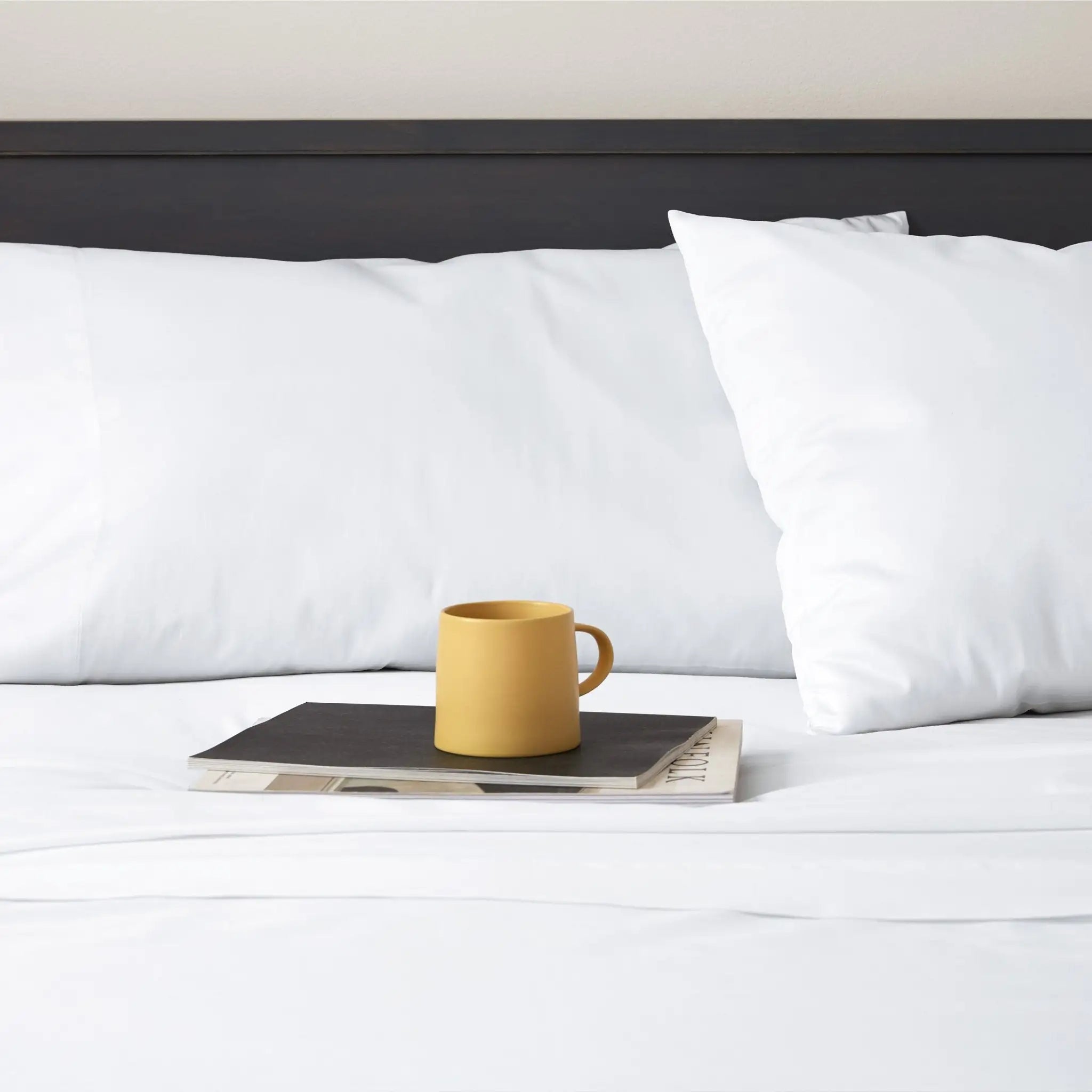 A yellow ceramic mug sits on a stack of magazines on a neatly made bed with white sheets and white pillows against a dark headboard.