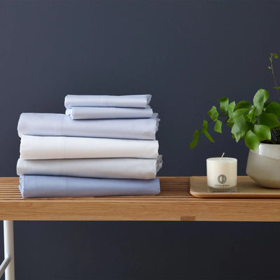 A stack of folded bedding in soft pastel shades of light blue, white, and pale gray sits on a wooden table.