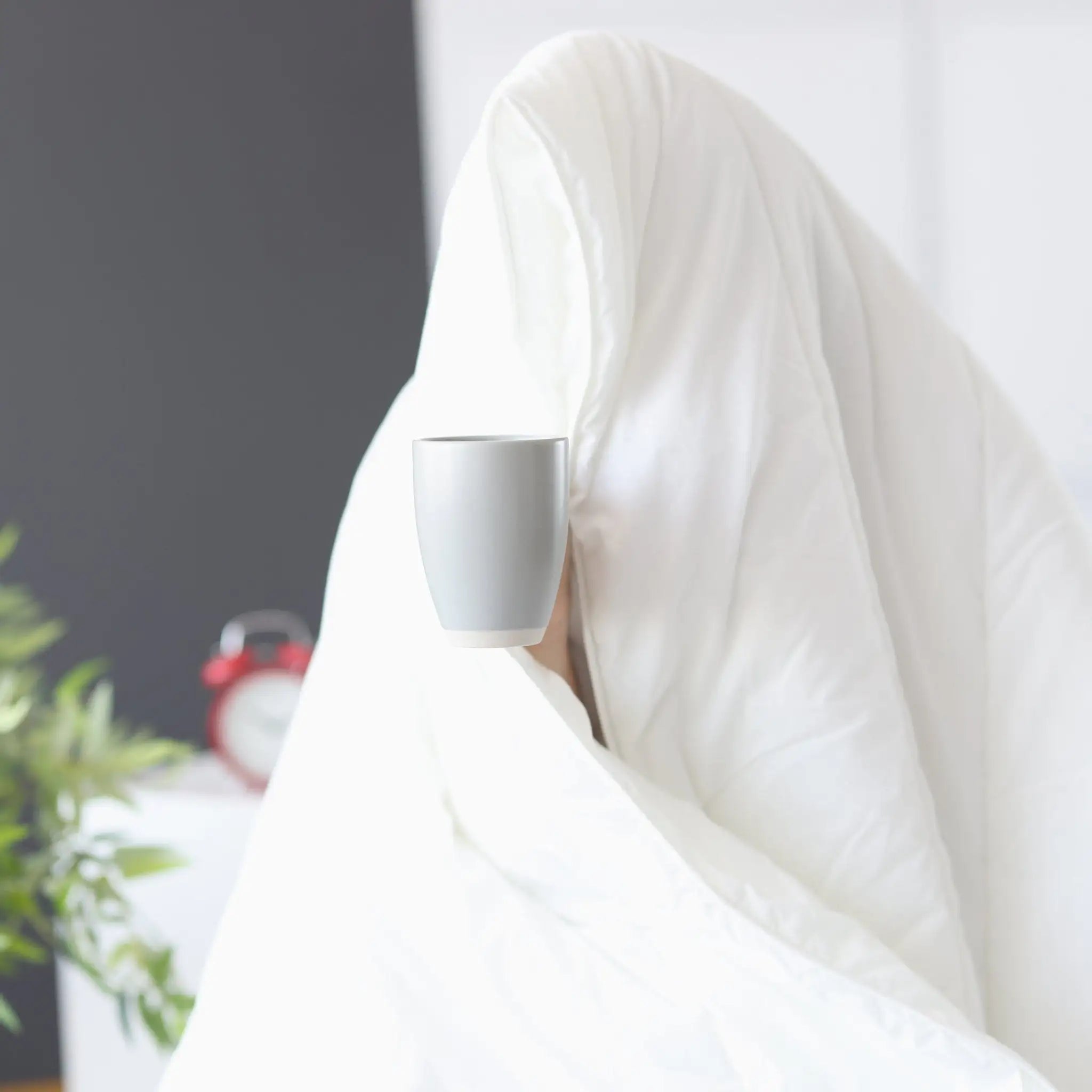 Person wrapped in a white comforter holding a white mug in a blurred indoor setting
