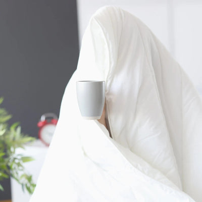 Person wrapped in a white comforter holding a white mug in a blurred indoor setting