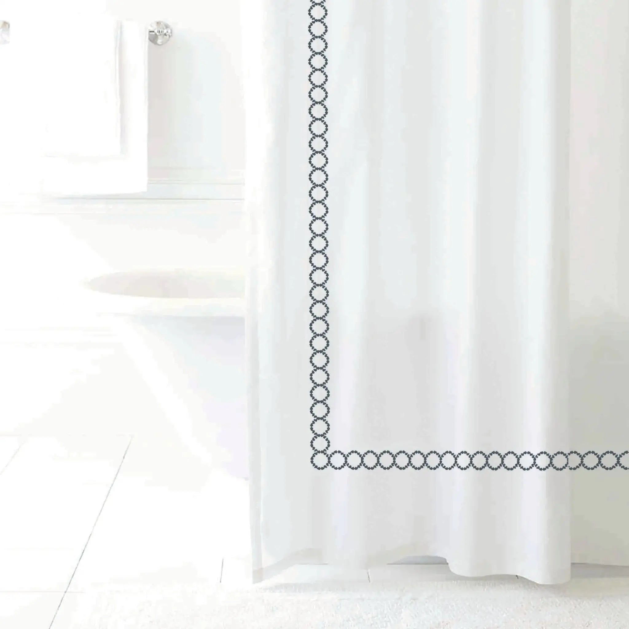 A white shower curtain with a charcoal chain-link border design, shown hanging in a bright, minimalist bathroom setting.