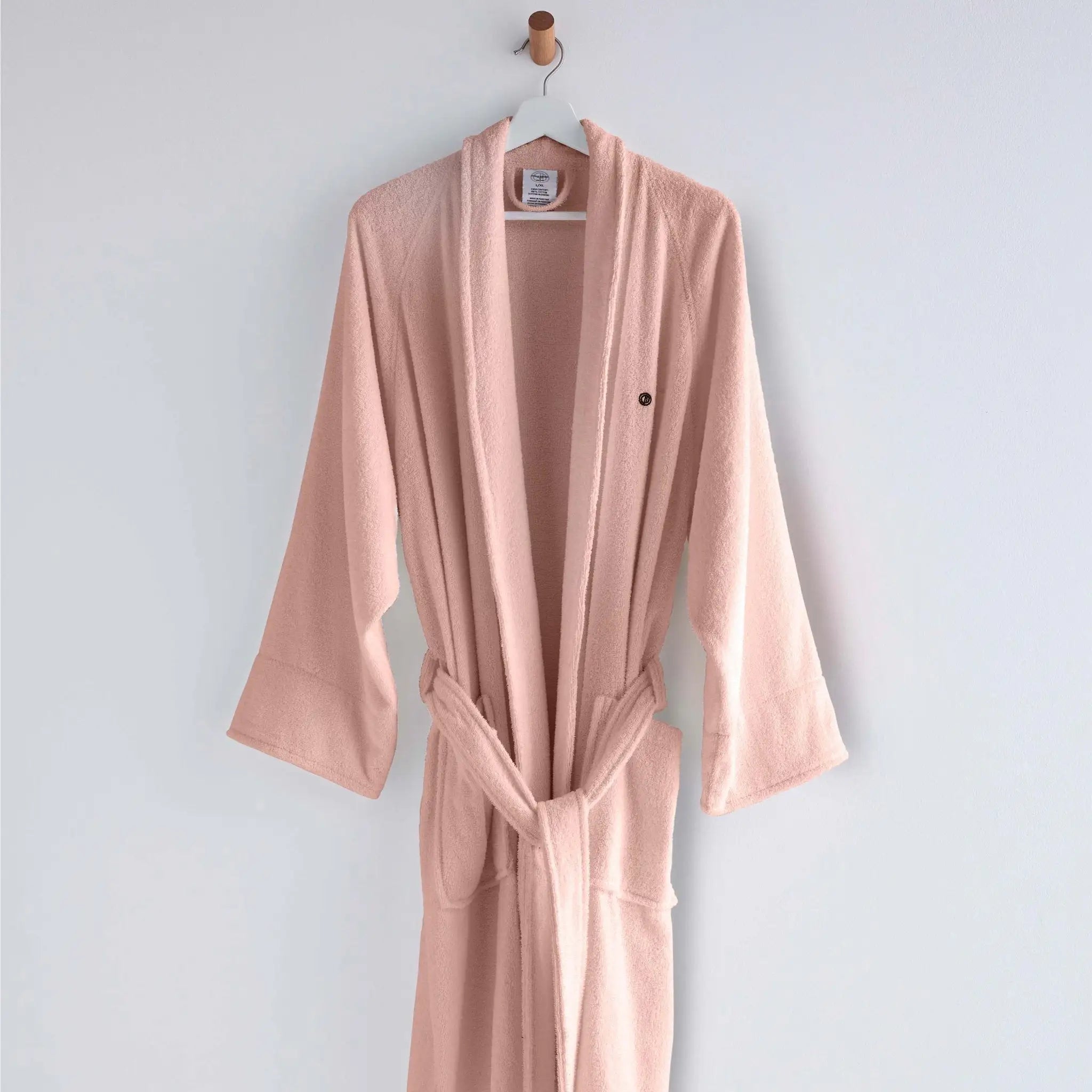 A light pink terry cloth bathrobe hanging on a wooden hanger against a plain light gray wall.