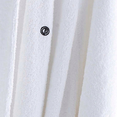 Close-up of a white textured fabric, likely a towel or bathrobe, with vertical folds and a small black circular embroidered logo near the center.