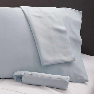Light blue bedding set including a pillowcase, folded pillowcase, and rolled sheet on a white bed against a dark gray headboard.