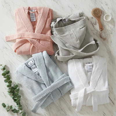 Four folded bathrobes in blush, gray, blue, and white are arranged on a white marble surface with a wooden brush, a small cup, and a green sprig.