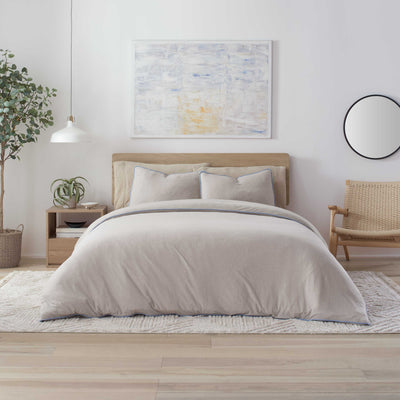 Light gray comforter set on a bed with matching pillows, surrounded by a plant, nightstand, and woven chair.