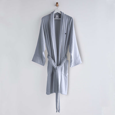 Light gray terry bath robe with long sleeves, a tie belt, and two front pockets, hanging on a wall.
