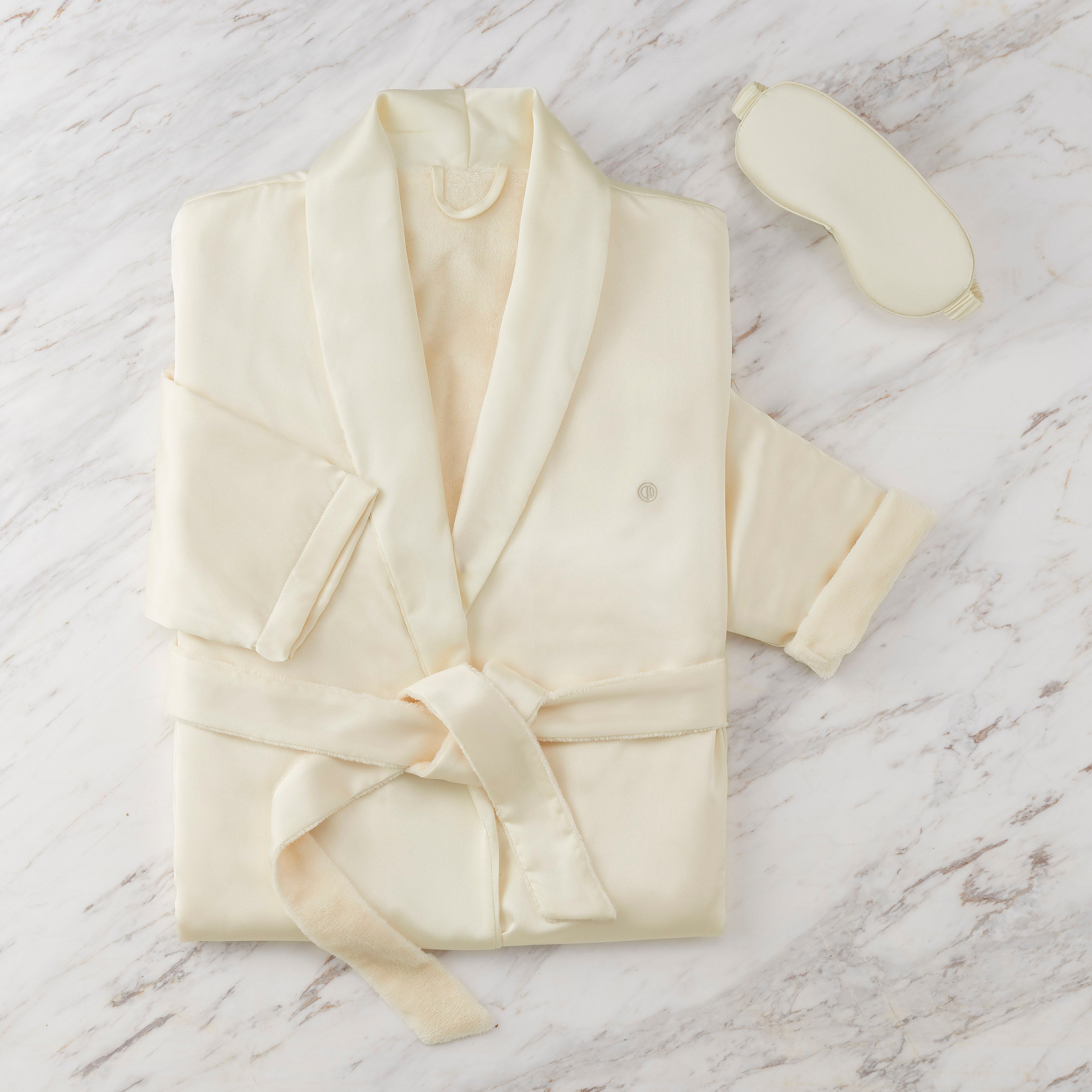 Satin robe in cream color with matching eyemask, laid flat on a marble surface.