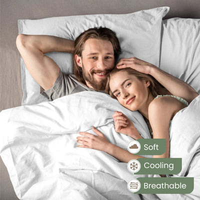 A smiling couple lying in bed under white bedding, with overlaid text badges reading Soft, Cooling, and Breathable. The scene is a real bedroom setting with pillows, a bed headboard, and the couple visible from head to upper torso.