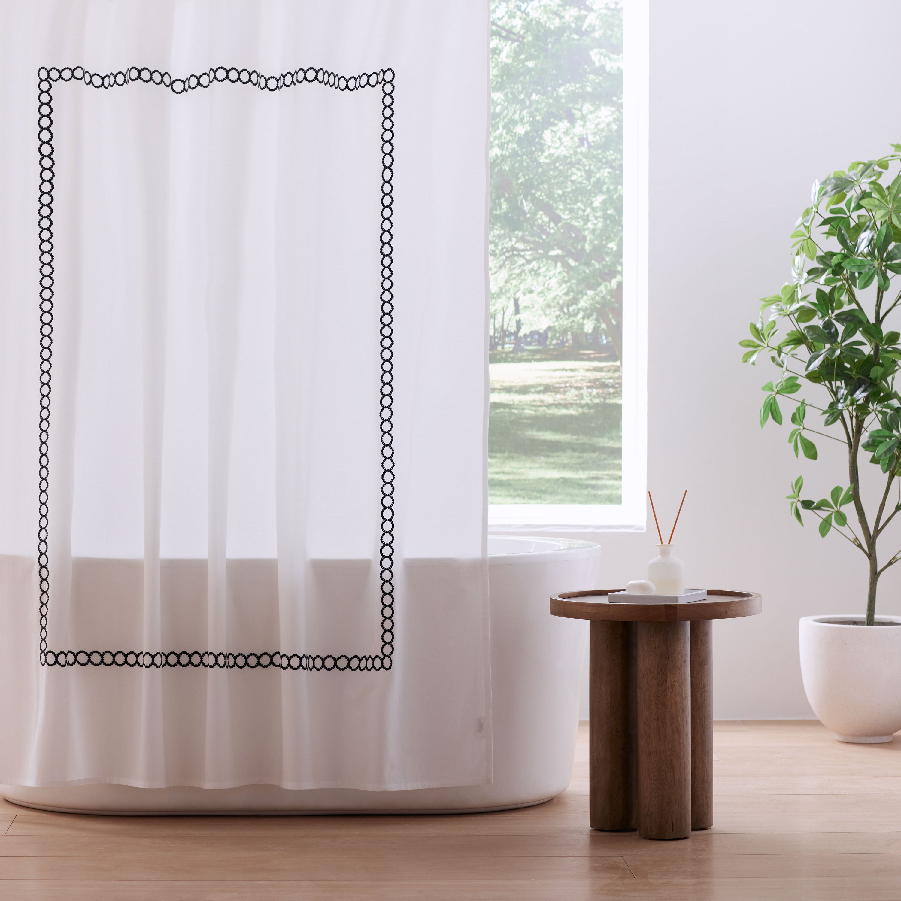 White shower curtain with a black circular border design, hanging near a bathtub and a wooden side table.