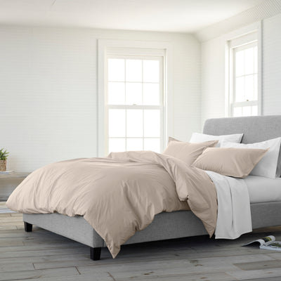 Beige duvet set on a gray upholstered bed with white and beige pillows in a bright, minimalist room.