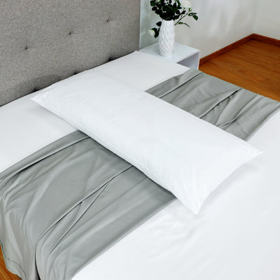 White gusset pillow placed on a bed with gray and white bedding, against a light-colored wall and a decorative vase.