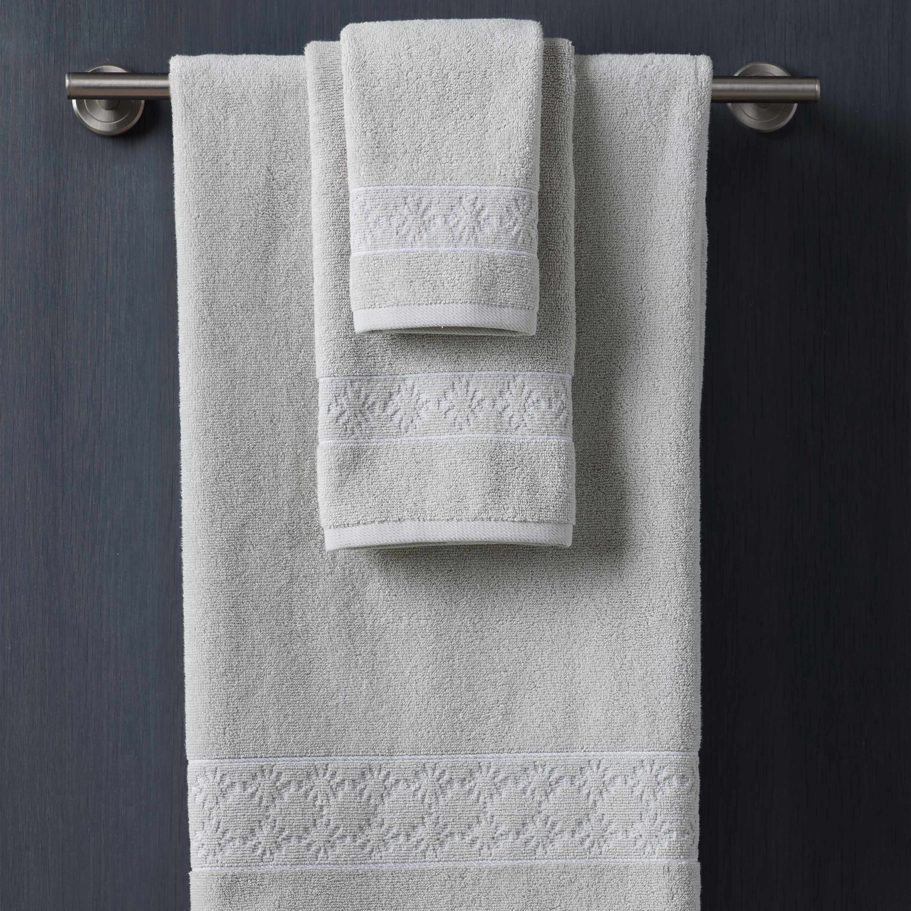 Three towels in light gray with decorative white embroidery, hanging on a silver towel bar against a dark background.