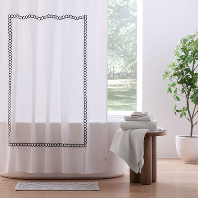 White shower curtain with a black circular border design, accompanied by stacked gray towels on a wooden stool.