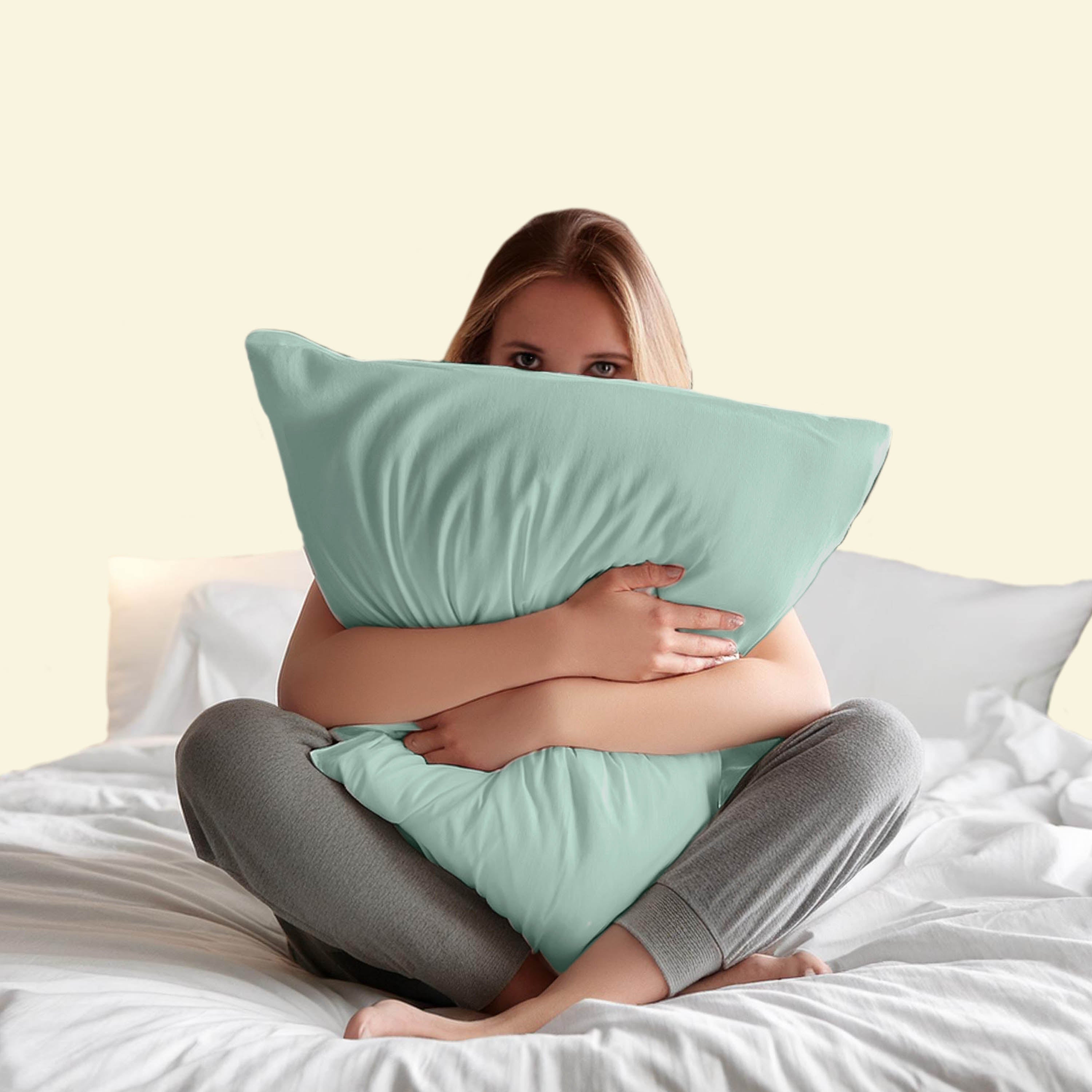 A person sitting on a bed holds two large mint green pillows while wearing gray pants.