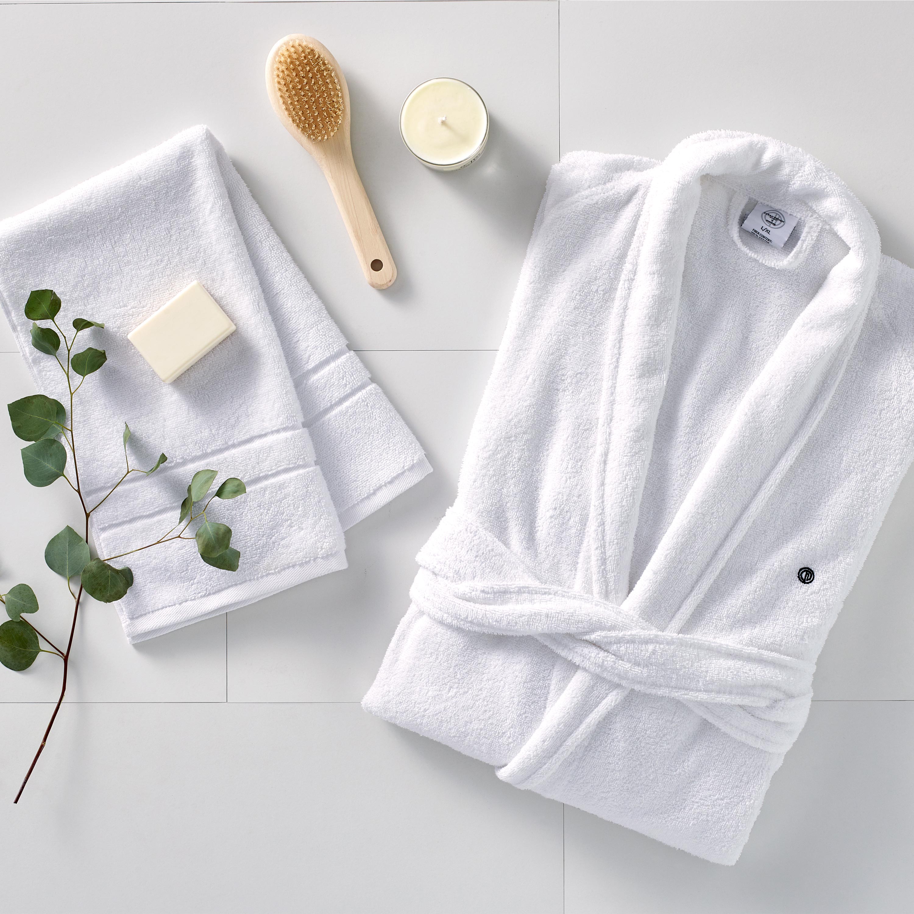 White terry bath robe with a tied waist, accompanied by two towels, a soap bar, a wooden brush, a candle, and a sprig of euca