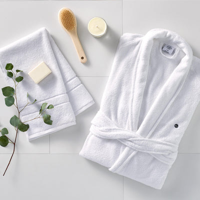 White terry bath robe with a tied waist, accompanied by two towels, a soap bar, a wooden brush, a candle, and a sprig of euca