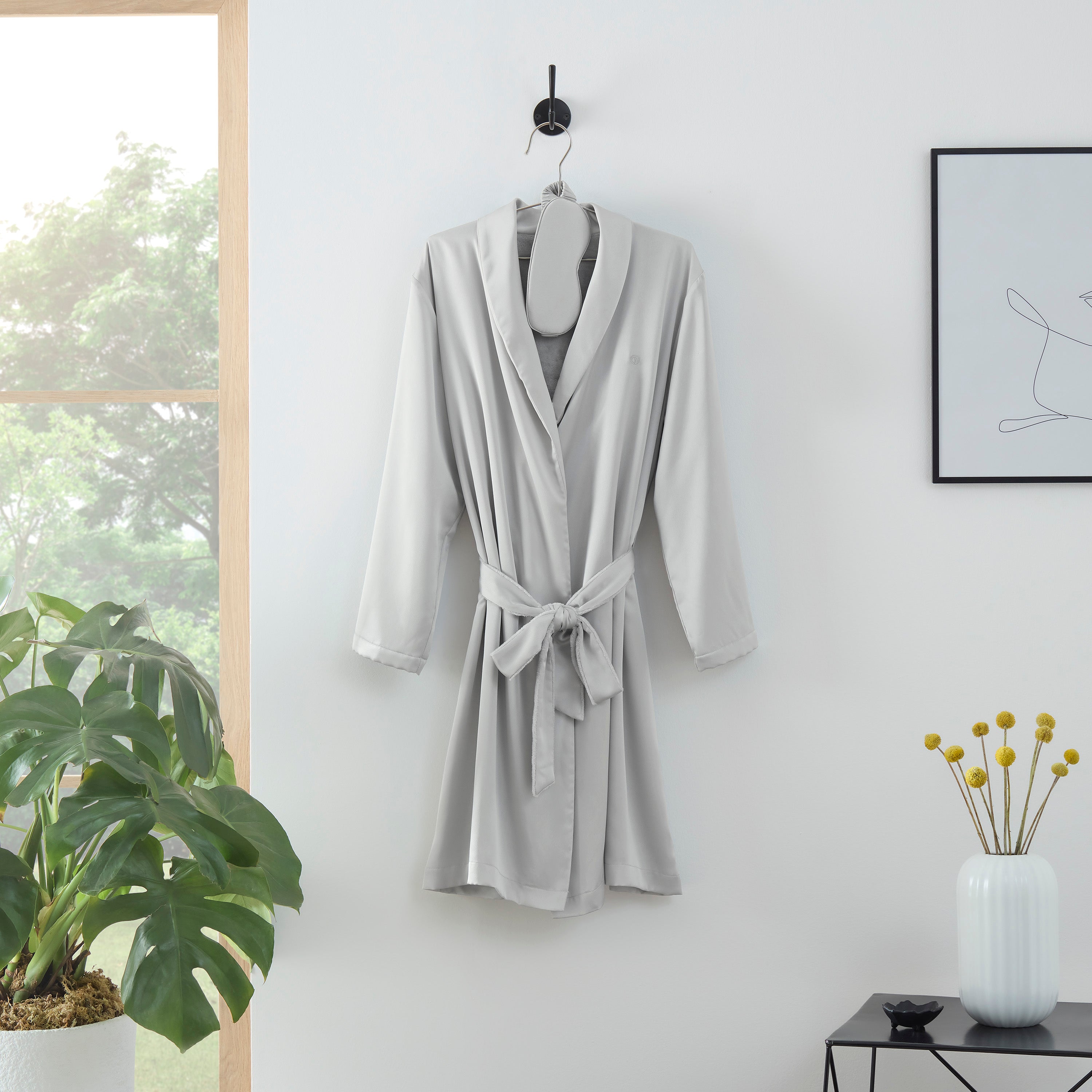 Satin robe with a tie belt and matching eyemask hanging on a hook against a light wall, with plants and decor nearby.