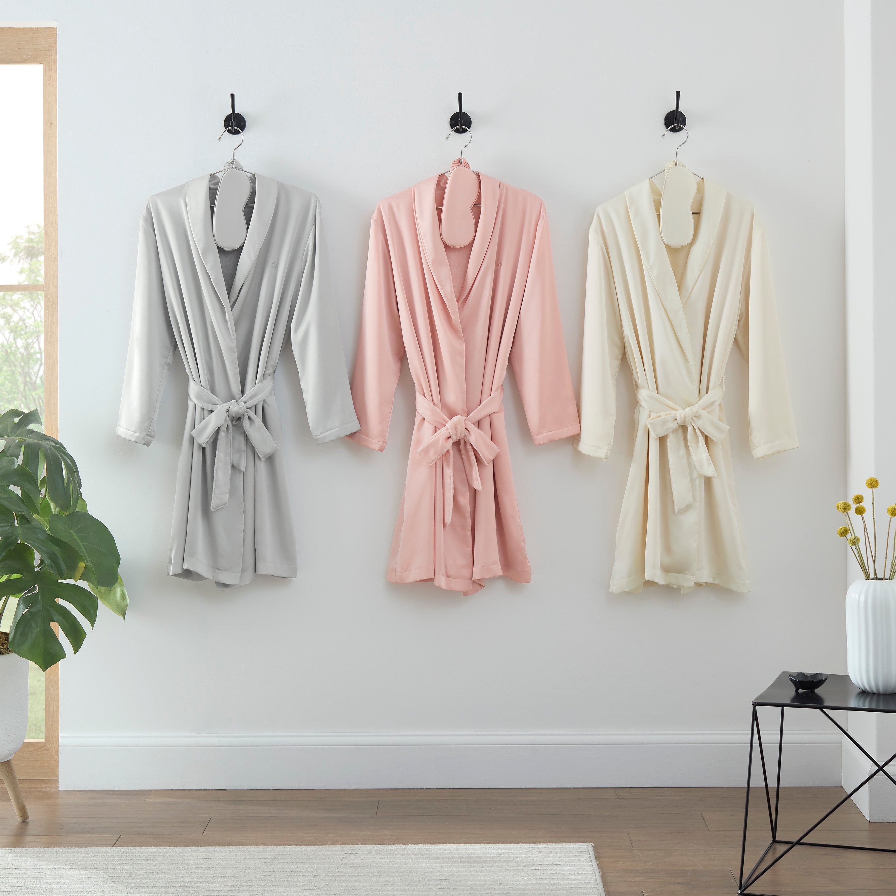 Three satin robes in gray, pink, and cream hanging on hooks, each with a matching eyemask.
