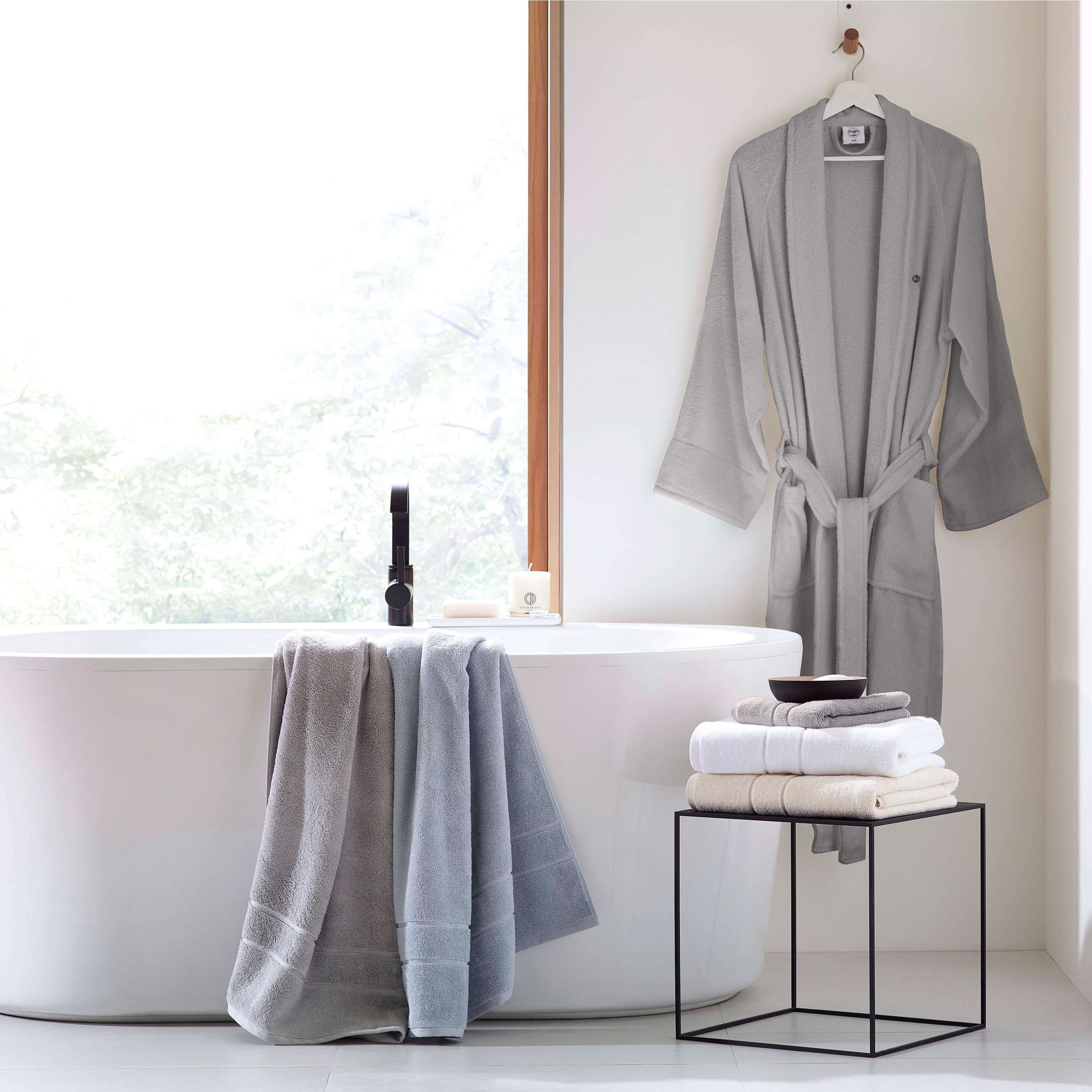 Gray terry bath robe hangs by a window, with towels draped over a bathtub and stacked on a black metal stand.
