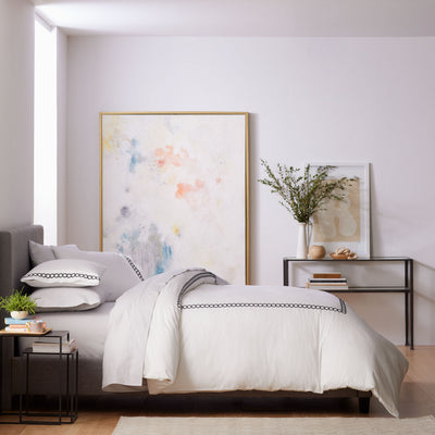Light-colored comforter set with decorative trim on a bed in a modern bedroom setting, featuring artwork and a side table.