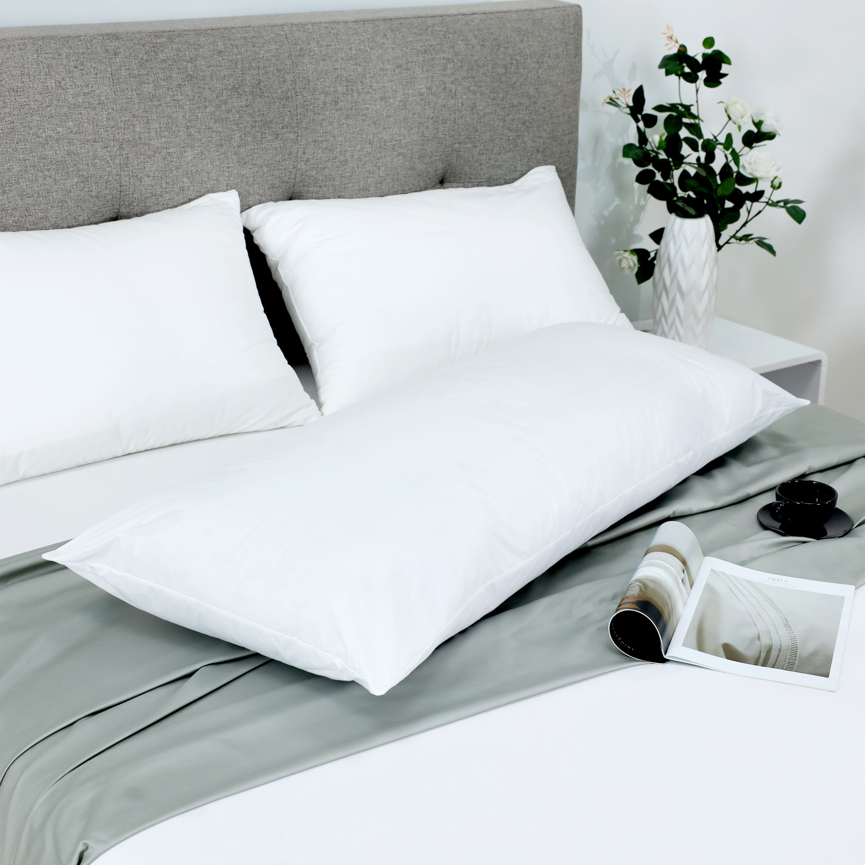 White gusset pillow on a bed with two additional white pillows, a green satin sheet, and a magazine beside a cup.