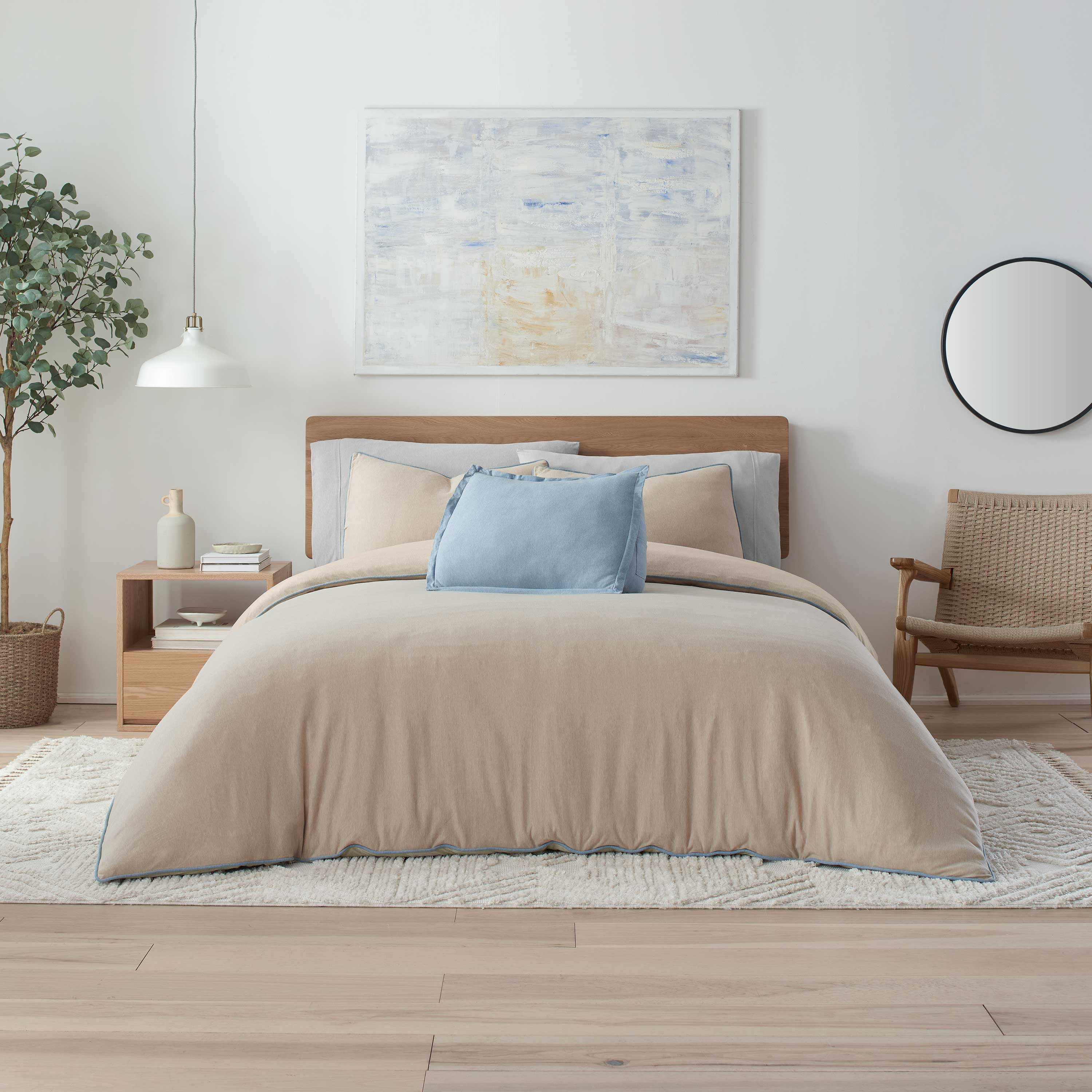 Beige and blue comforter set on a bed with decorative pillows, surrounded by a plant, nightstand, and chair in a bright room.