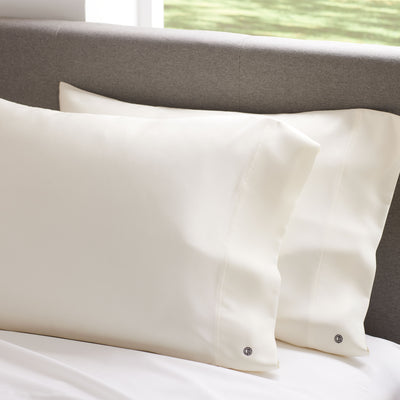 Two cream satin standard pillowcases resting on a white bedspread against a gray headboard.