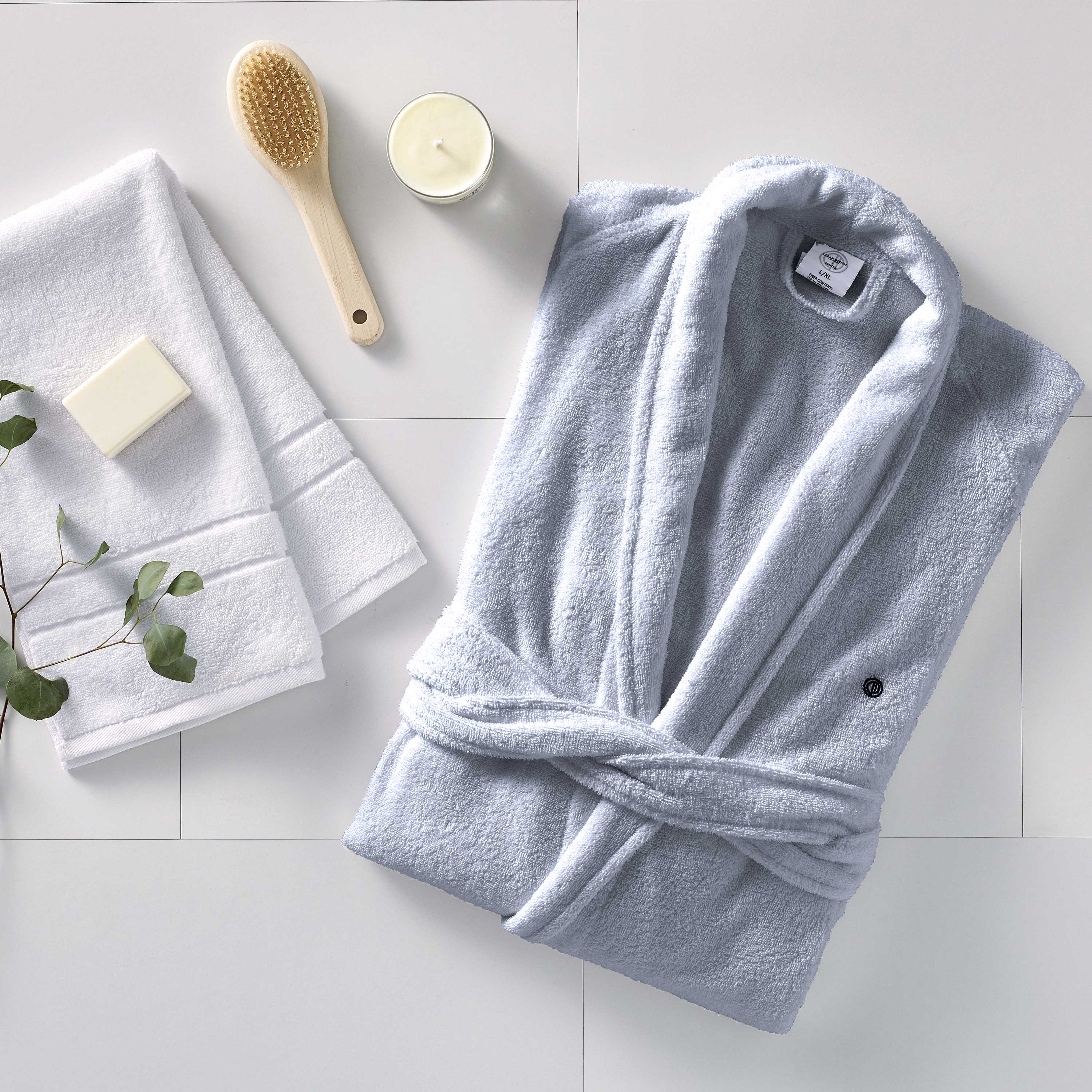 Light blue terry bath robe with a tied waist, accompanied by white towels, a soap bar, a wooden brush, and a candle.