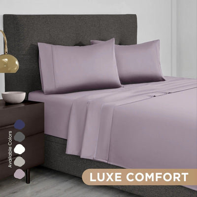A bed dressed in a lilac bedding set with two pillowcases and a matching sheet, shown in a styled bedroom scene with a dark upholstered headboard, bedside lamp, and a text badge reading 'LUXE COMFORT' plus an 'Available Colors' swatch panel.