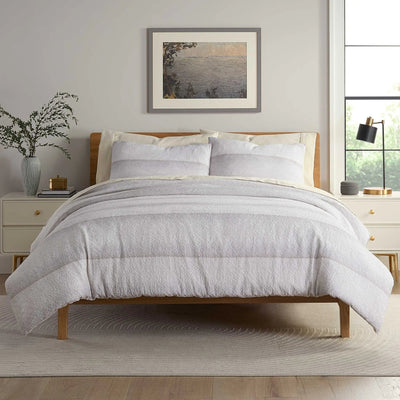 A neatly made bed with a light gray textured comforter and matching pillow shams, set in a bright bedroom.