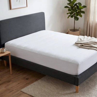 A white quilted mattress topper or mattress protector shown on a dark gray upholstered bed frame in a styled bedroom setting.