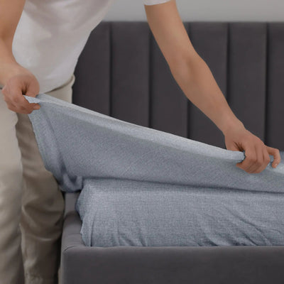 A person stretching a light blue-gray fitted sheet over a mattress on a gray upholstered bed, shown in a clean indoor setting.