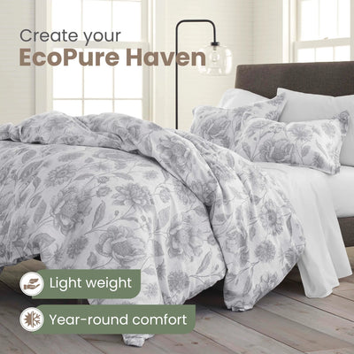 A bed dressed in a light gray and white floral-patterned comforter and matching pillowcases, shown in a bright bedroom setting with text overlays reading 'Create your EcoPure Haven' and 'Light weight Year-round comfort'.
