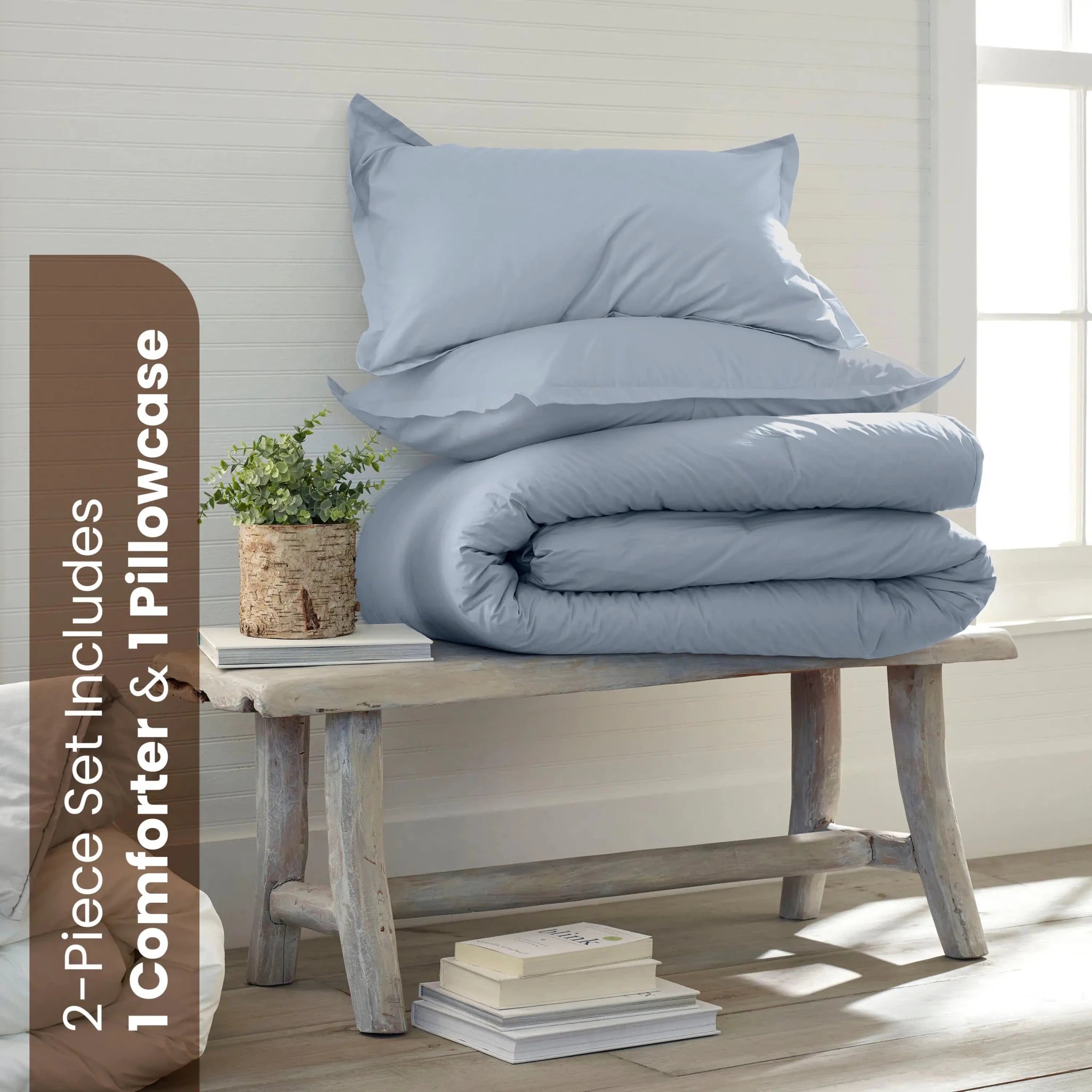 A bedding set with a light blue comforter and two matching shams displayed on a rustic wooden bench in a bright room. A brown text banner on the left reads '2-Piece Set Includes 1 Comforter & 1 shams'.
