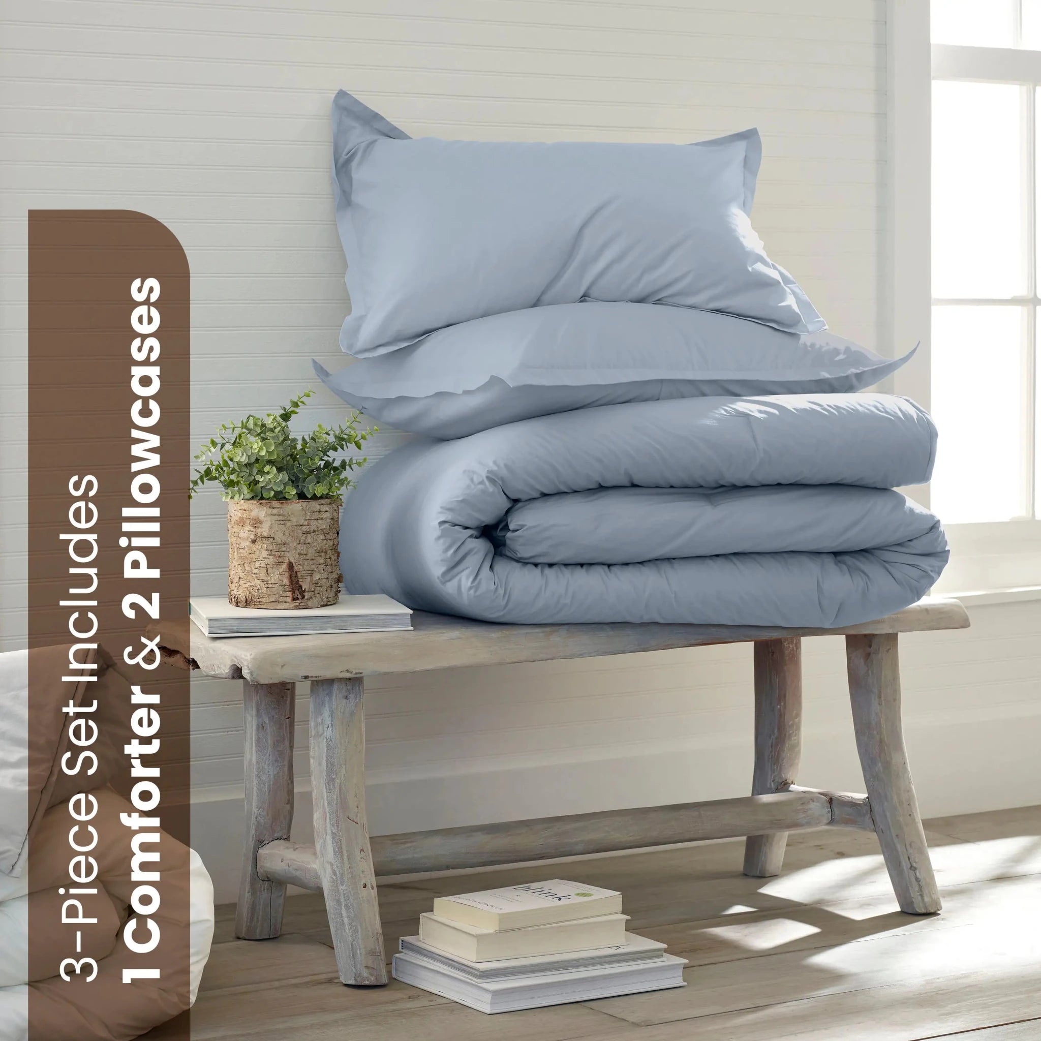A light blue bedding set consisting of a comforter and two shams, neatly stacked on a rustic wooden bench in a bright, sunlit room. A brown vertical banner on the left reads '3-Piece Set Includes 1 Comforter & 2 shams'.