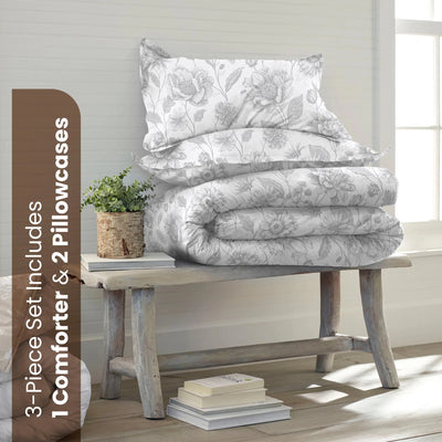 A bedding set displayed on a rustic wooden bench in a bright room. The set includes a white comforter and two matching pillowcase with a gray floral pattern.
