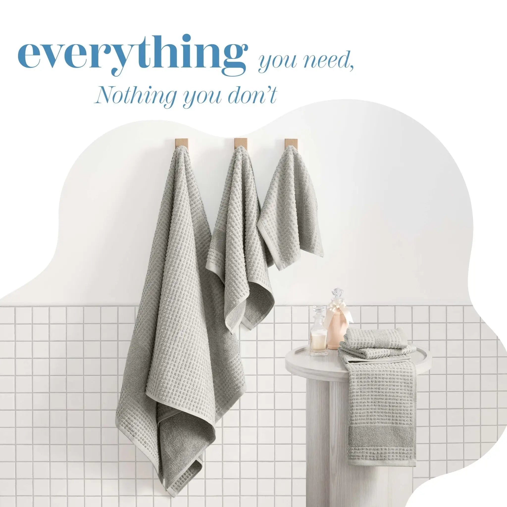 Three light gray towels hang from wooden pegs on a white tiled wall and a decorative text overlay at the top reading 'everything you need, Nothing you don't'.