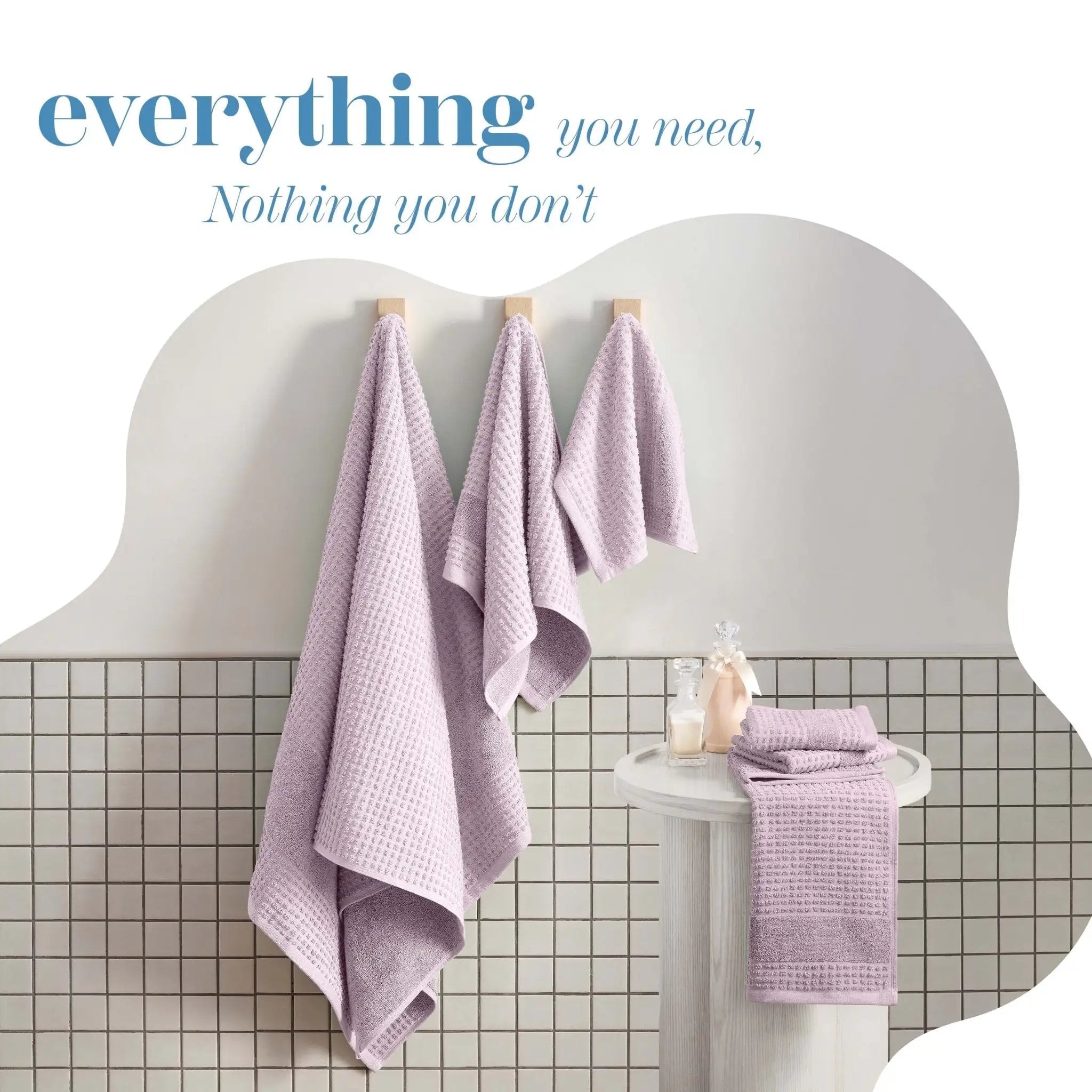 Three liliac towels hang on wooden hooks against a white wall above a tiled pedestal, with the text 'everything you need, Nothing you don't' at the top.