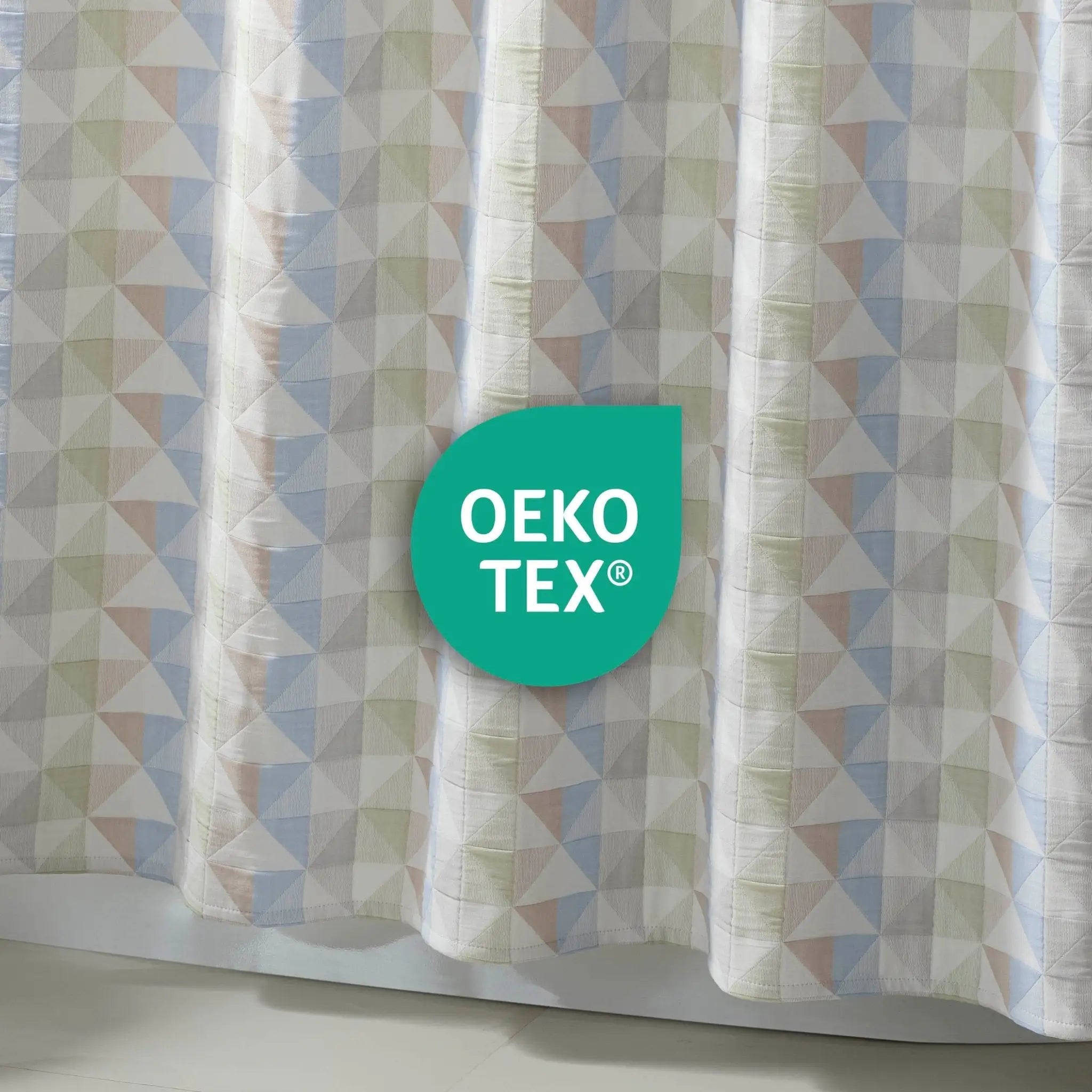 A close-up of a patterned curtain with a geometric triangle design in white, light blue, and pale green. A teal circular badge in the center reads 'OEKO TEX™'.