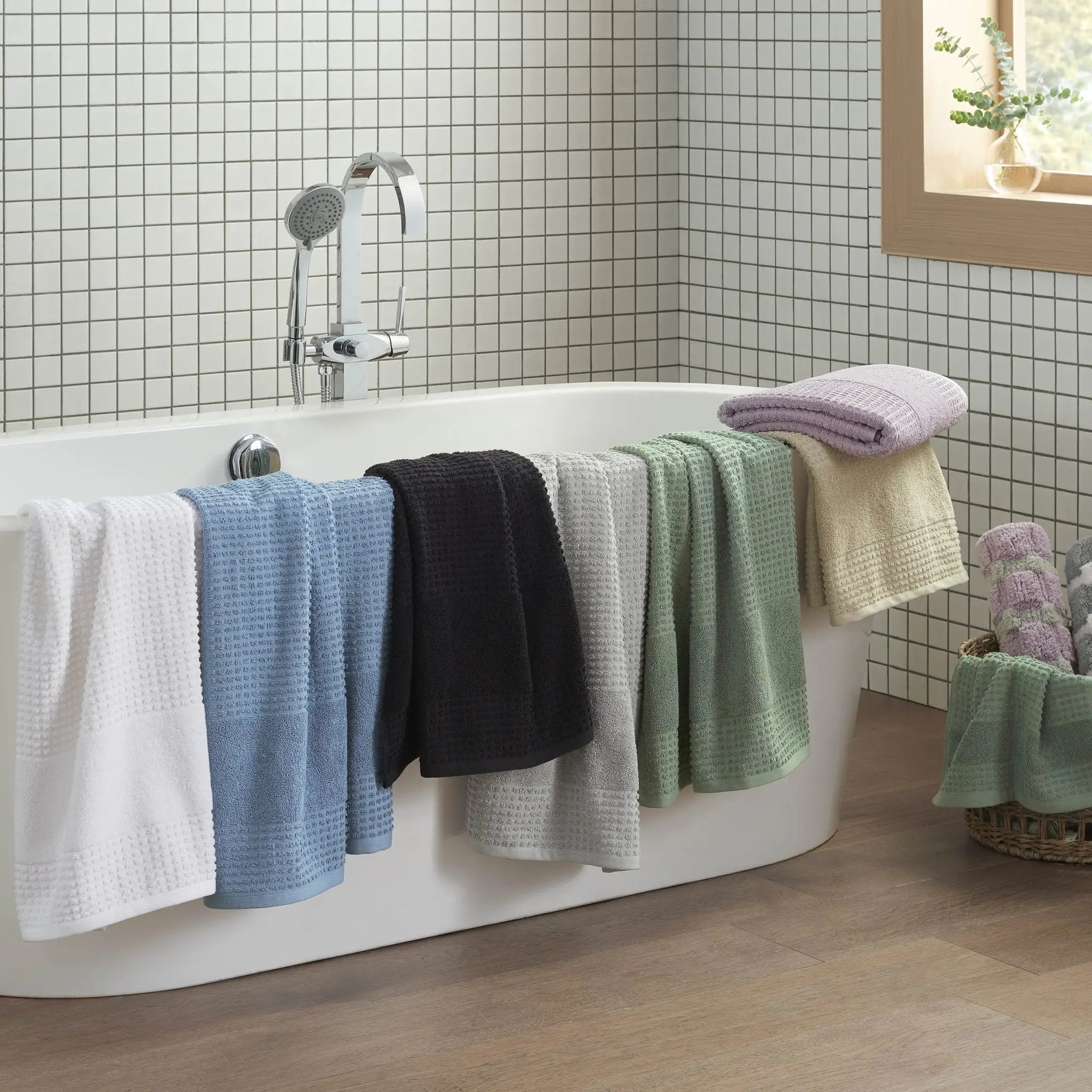 A set of textured bath towels in white, light blue, black, light gray, sage, cream, and lavender, draped over the side of a white freestanding bathtub in a tiled bathroom.