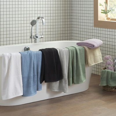 A set of textured bath towels in white, light blue, black, light gray, sage, cream, and lavender, draped over the side of a white freestanding bathtub in a tiled bathroom.