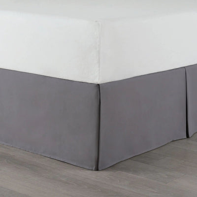 A gray bed skirt made of fabric, shown on a bed with a white fitted sheet and gray wood-look flooring.