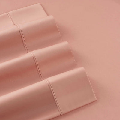 A close-up of folded dusty pink bedding, likely a pillowcase or sheet set, arranged diagonally on a matching solid pink background.