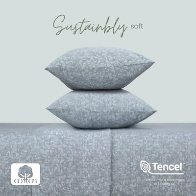Two light gray textured pillows stacked on a matching gray upholstered surface, with the words 'Sustainably soft' above. Cotton and Tencel logos appear at the bottom, with a pale gray background.