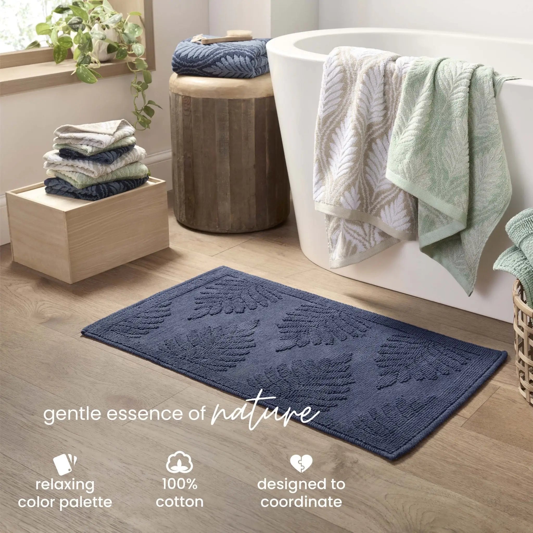 A navy-blue bath rug with a raised leaf pattern is shown in a bright bathroom setting next to a white freestanding tub, with folded towels. Text at the bottom reads 'gentle essence of nature' and includes feature callouts.