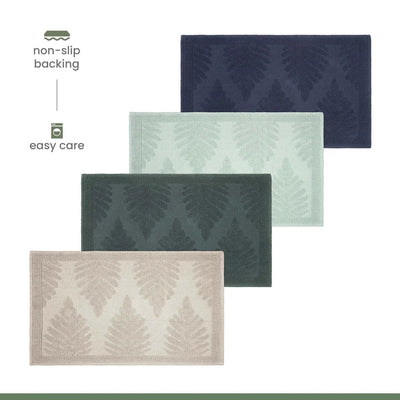 Four rectangular bath mats with a raised fern pattern are shown on a white background, arranged diagonally. The mats come in navy, sage, jade, and sand.