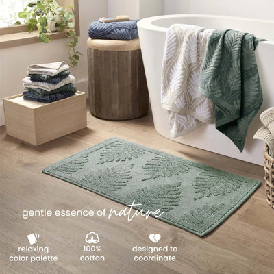 A sage bath rug on a light wood floor in a bathroom setting, with a white freestanding tub, folded towels, and decorative text at the bottom.