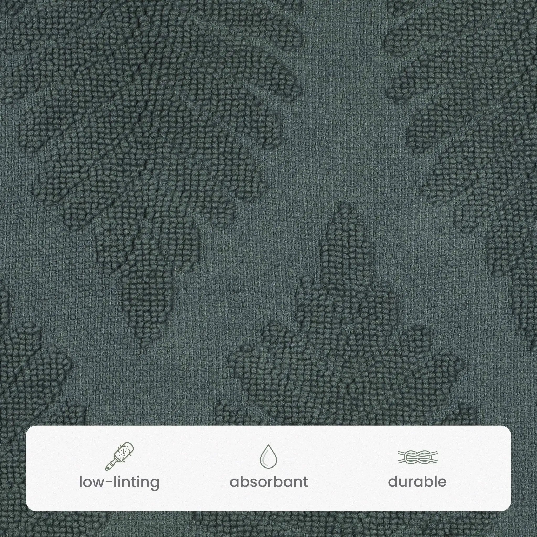 Close-up of a jade bath rug textured fabric with a subtle tonal leaf pattern. A white label at the bottom lists features: low-linting, absorbent, and durable.